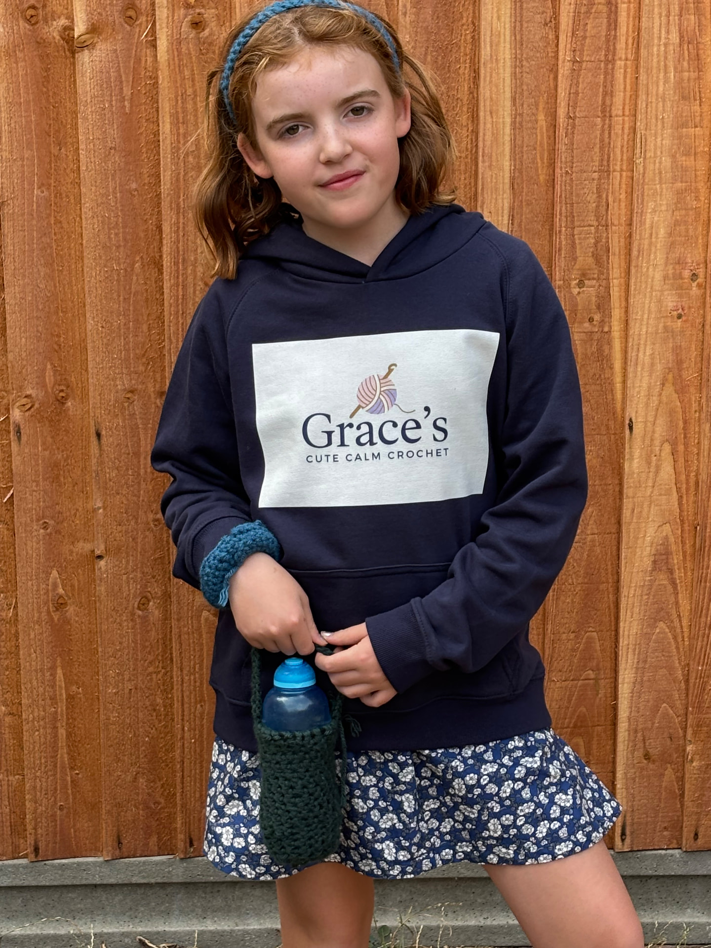 Girl wearing a crochet headband and Grace's Cute Calm Crochet hoodie, carrying a green crochet bottle carrier