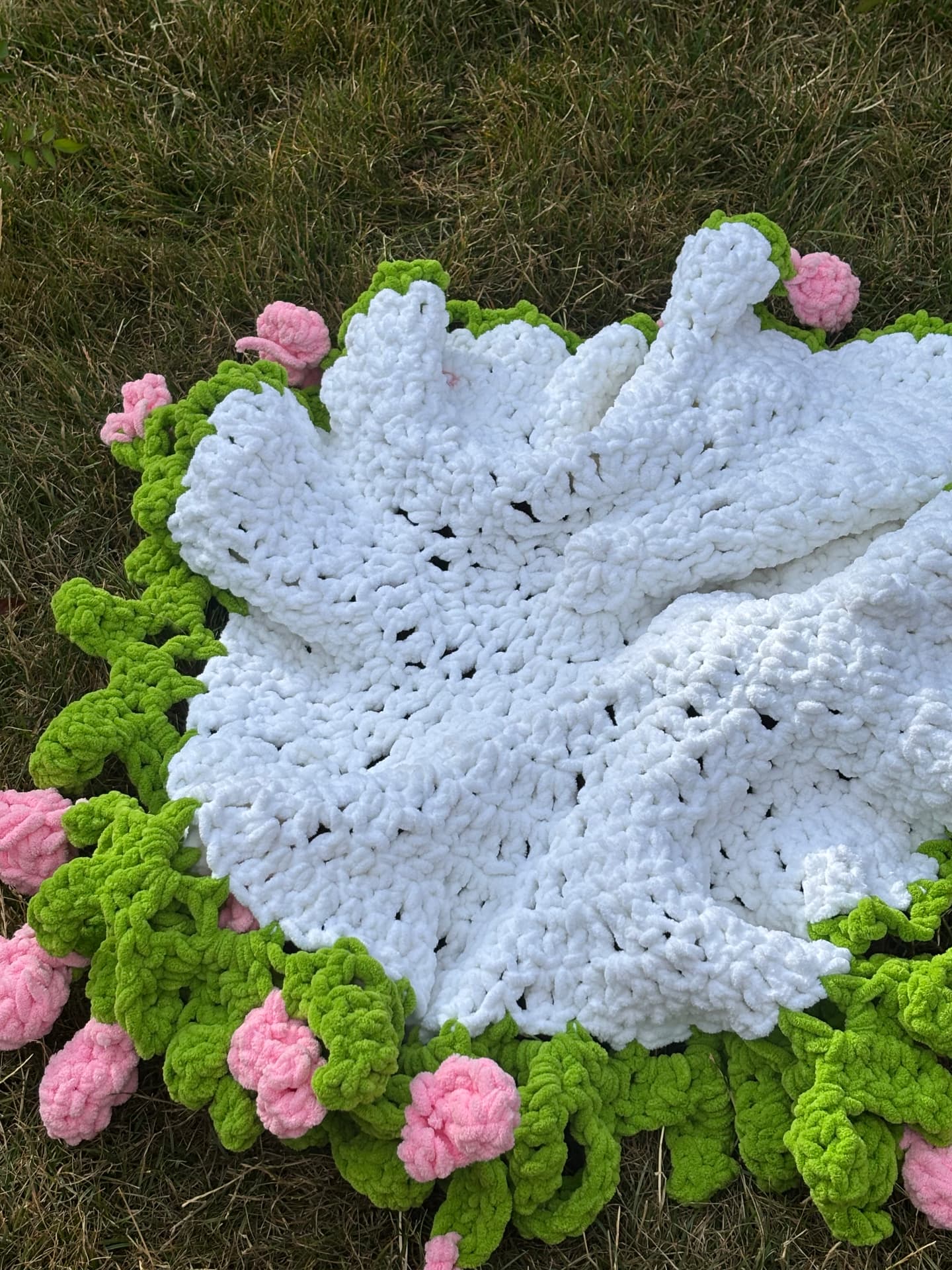 Chunky white crochet blanket with green leaf and pink rose border laid on grass