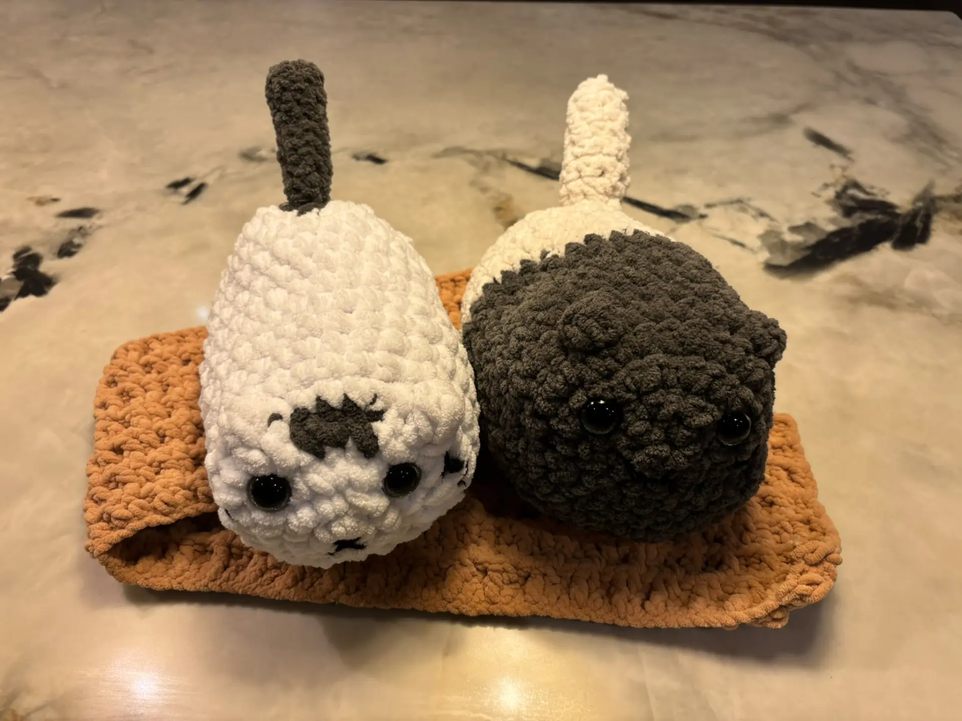 Two chunky crochet loaf cats, one white with dark markings and one dark grey, sitting side by side on a tan crochet mat